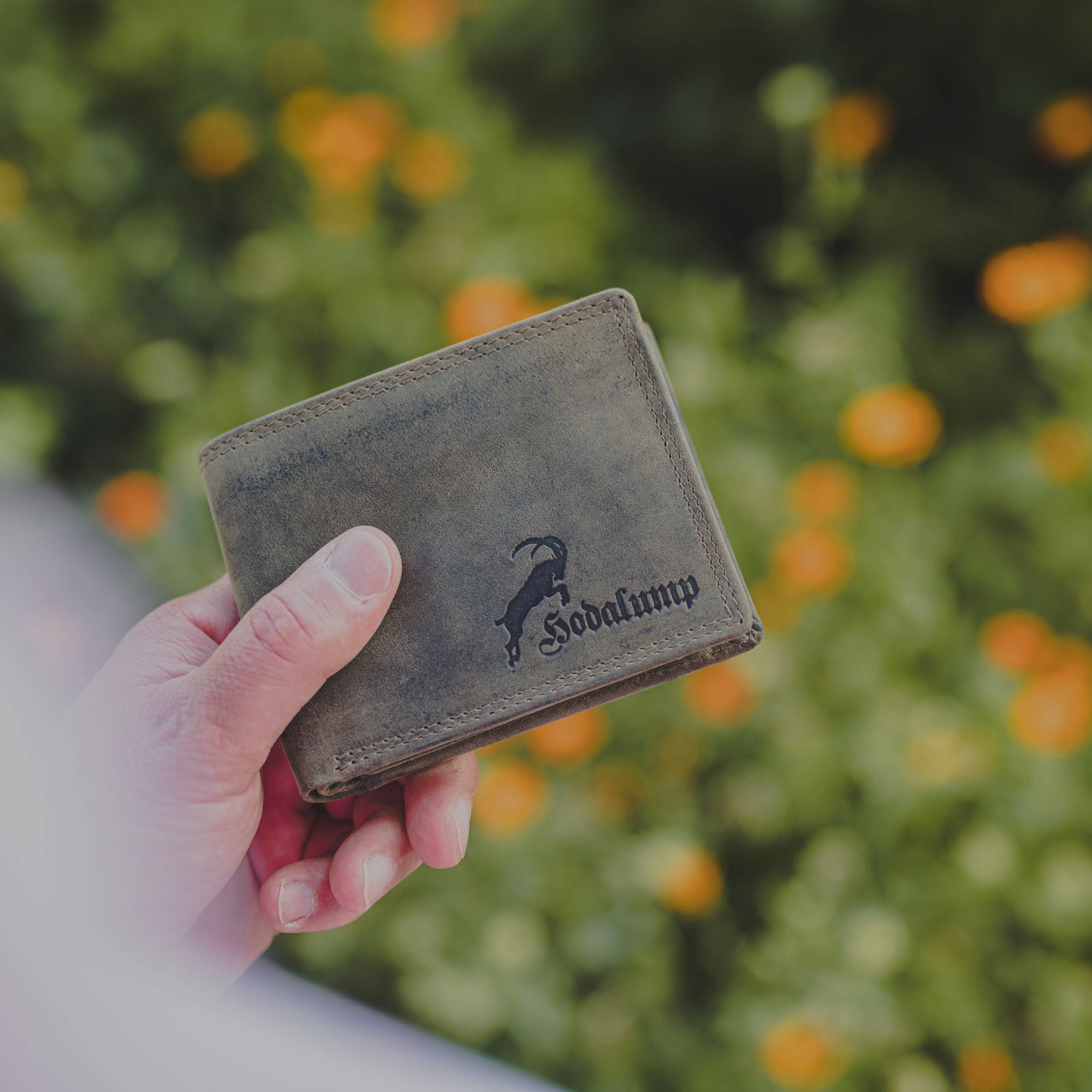 Hand hält eine braune Leder-Geldbörse mit schwarzem Logo und Schriftzug ‚Hodalump‘ vor einem unscharfen Hintergrund mit grünen Pflanzen und orangefarbenen Blumen